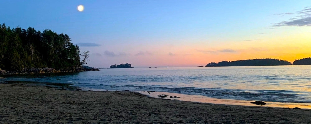Tranquil beach at sunset with a full moon rising over calm waters and silhouetted islands in the distance.