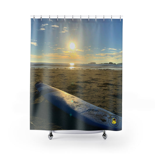 Sunset over a sandy beach with a surfboard in the foreground and soft clouds glowing above the ocean horizon.